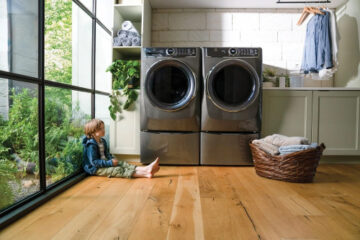 An electrolux dryer and washer in a luxury home. A small boy sits next to the appliances on hardwood floors.