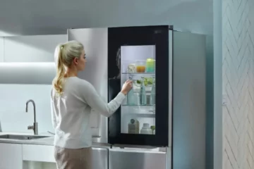 A young woman with blonde hair tied back in a ponytail stands in front of a sleek, high-tech LG refrigerator. She's wearing a light-colored, long-sleeved top and is reaching out to touch the refrigerator's door.