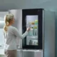 A young woman with blonde hair tied back in a ponytail stands in front of a sleek, high-tech LG refrigerator. She's wearing a light-colored, long-sleeved top and is reaching out to touch the refrigerator's door.
