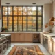 Head-on shot of an inviting cozy kitchen. Pumpkin pie on the counter, the space features warm lighting and cream walls with wide oak cabinets. There is an island in front of large windows where autumn leaves can be seen. Luxury appliances visible, including upscale fridge and wine cooler.