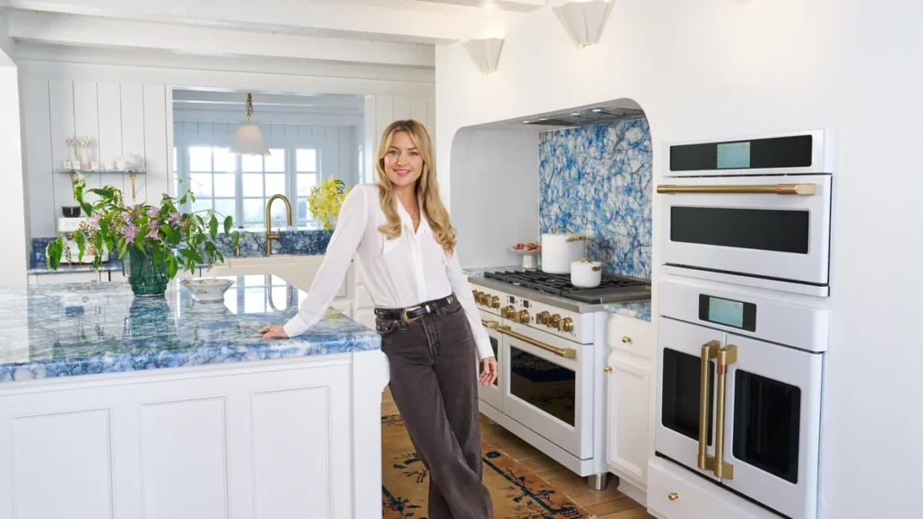 Kate Hudson standing in her blue jean baby kitchen with Sodalite Blue Quartzite countertops and Cafe Matte White appliances with Brushed Brass hardware